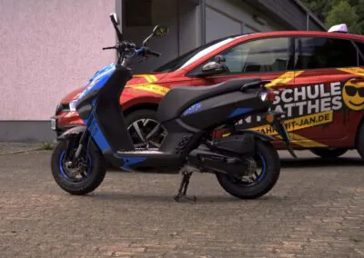 Blauer Motorroller vor dem Fahrschulauto der Fahrschule Jan Matthes