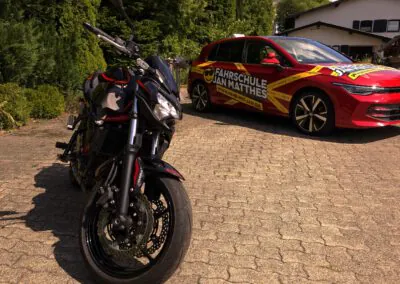 Schwarzes Kawasaki Z650 Motorrad im Vordergrund und rotes VW Golf 8 Fahrschulauto der Fahrschule Jan Matthes in Bad Berleburg