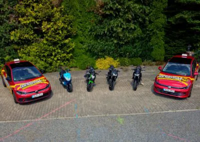 Luftaufnahme des gesamten Fuhrparks der Fahrschule Jan Matthes in Bad Berleburg mit zwei PKW, einem Roller und mehreren Motorrädern auf dem Parkplatz